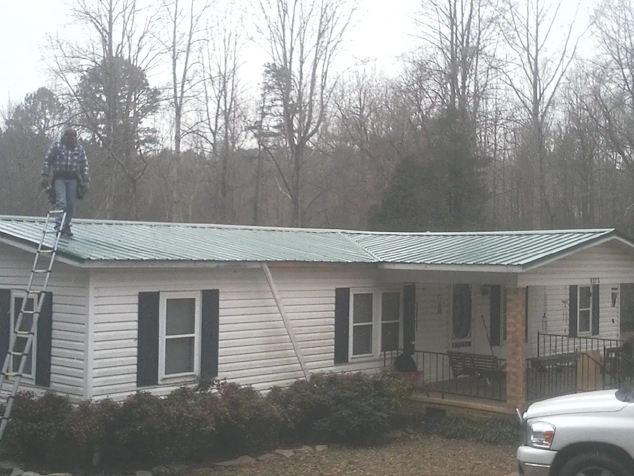 Worker on a ladder beside a new green metal roof on a ranch home in Cartwright