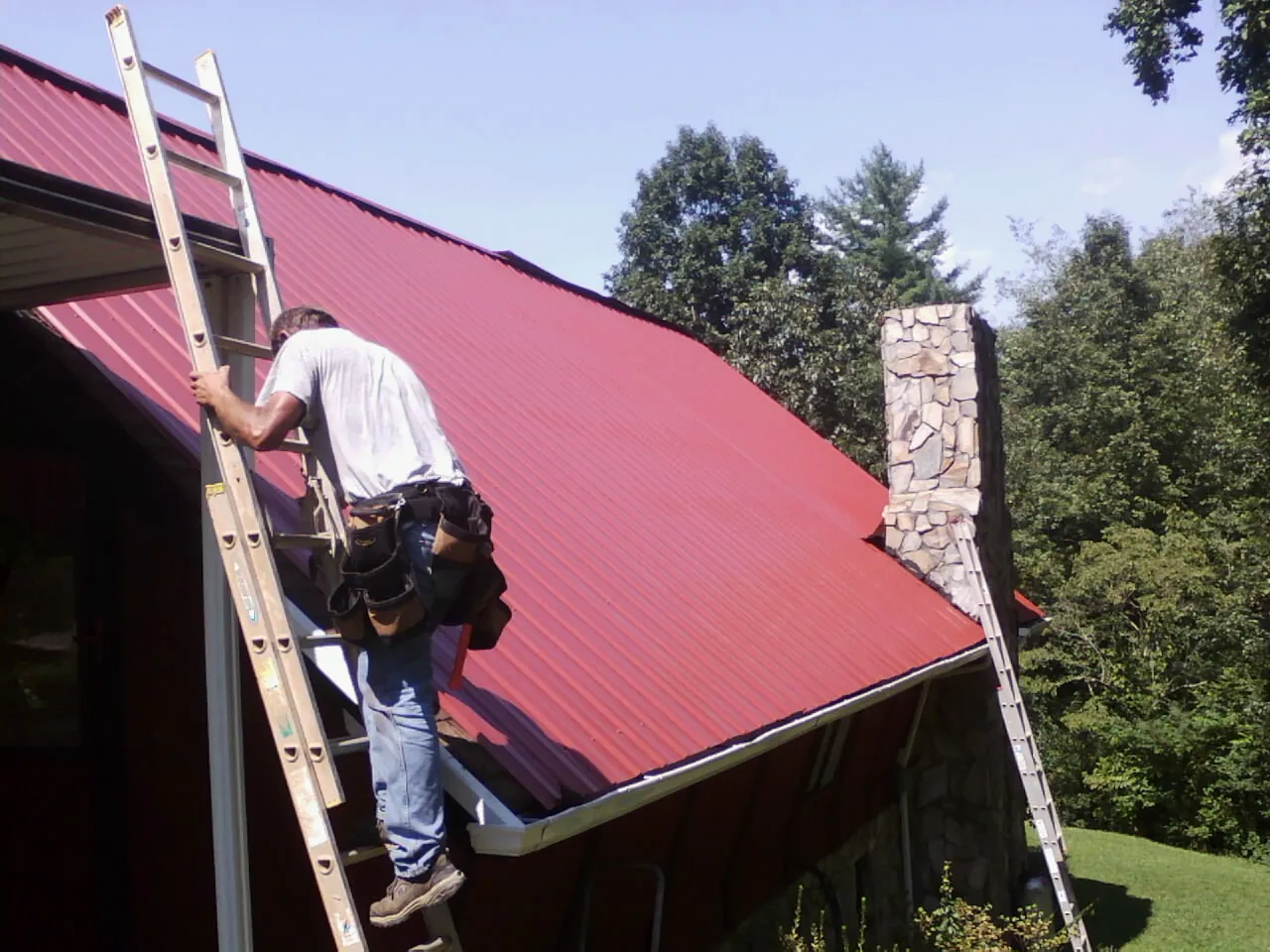 Roofer on a ladder beside a red metal roof and stone chimney — Soffit & Fascia Repair by Roofing Brothers in Cartwright